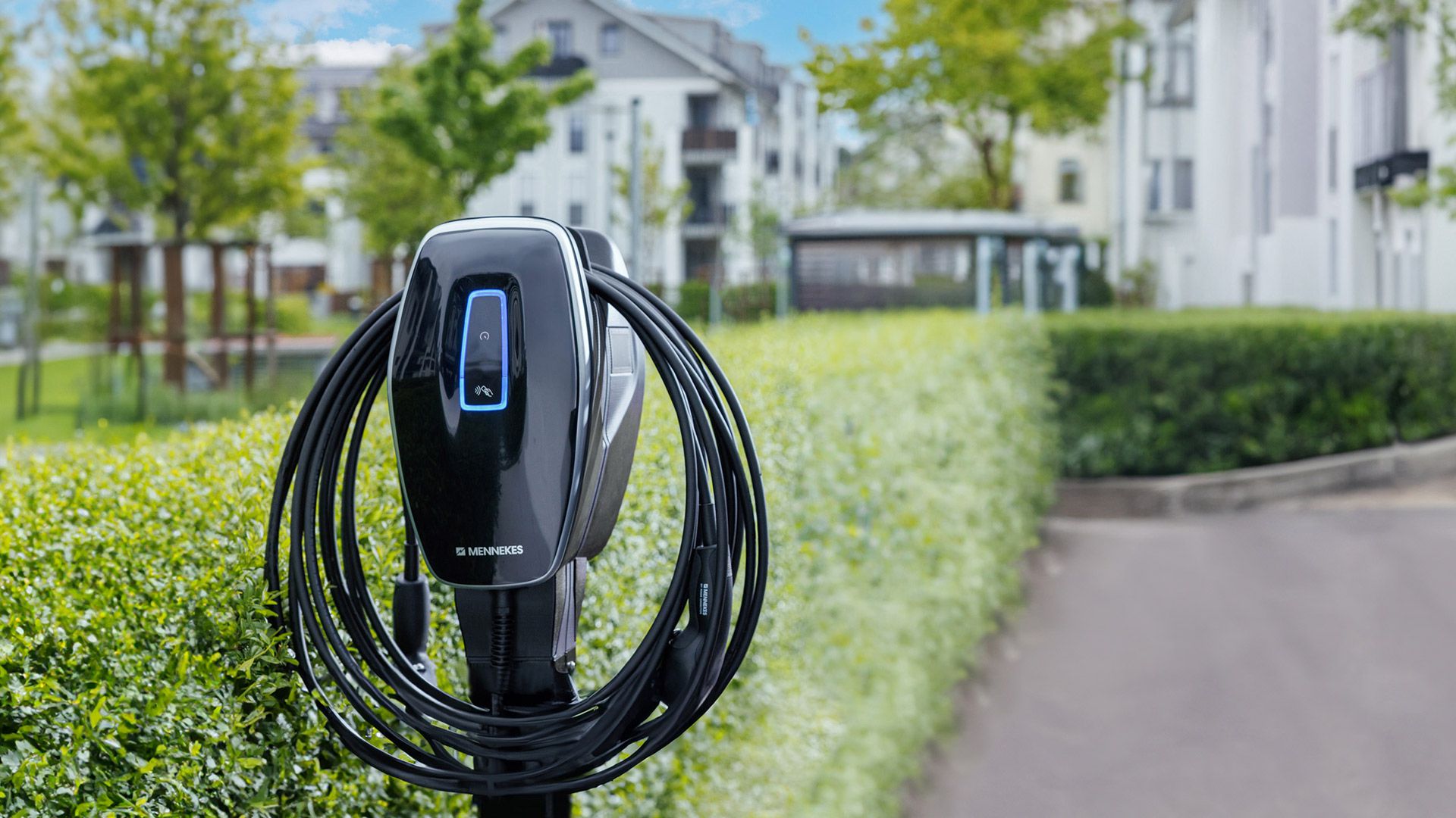Förderung von Elektromobilität Ladestation von Mennekes mit Ladekabel im Außenbereich vor modernen Wohn- oder Bürogebäuden und grüner Hecke.